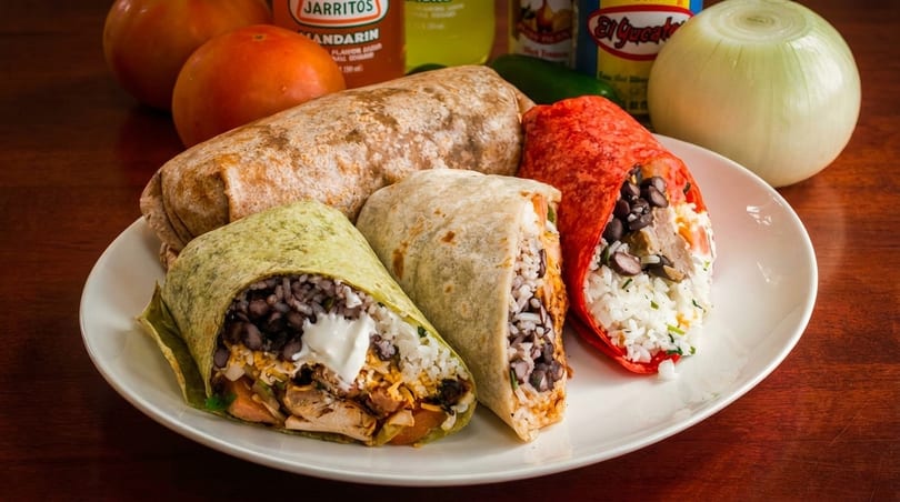 A plate of three colorful burritos, one cut open, with fresh ingredients and drinks in the background.