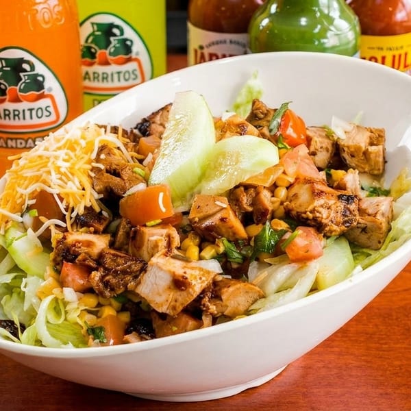 A white bowl filled with a chicken salad, topped with cheese, tomatoes, corn, and cucumbers.