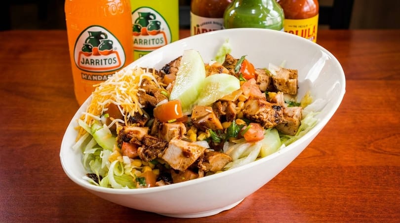 A white bowl filled with a chicken salad, topped with cheese, tomatoes, corn, and cucumbers.