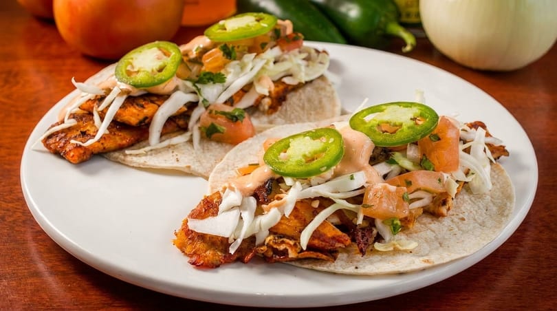 Two grilled chicken tacos with cabbage, salsa, sauce, and jalapeños on a white plate.