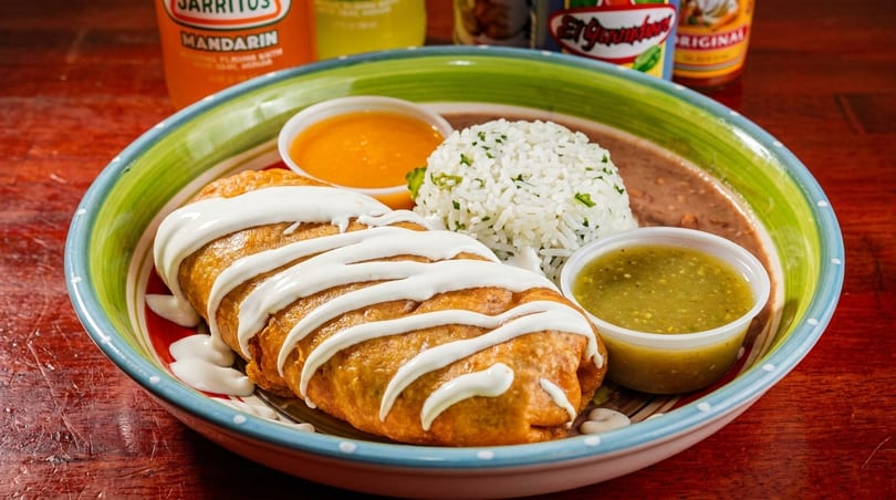 A deep-fried burrito drizzled with sour cream, served with rice, refried beans, and two dipping sauces.