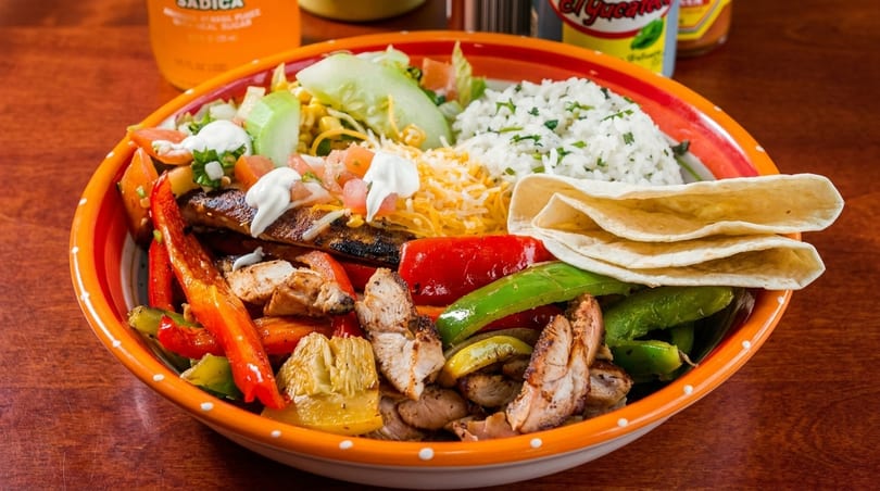 A colorful fajita bowl with grilled chicken, peppers, onions, rice, salad, cheese, and tortillas.